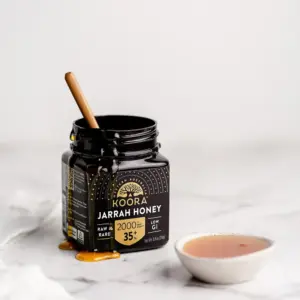 Open jar of Koora Jarrah Honey TA35+ with a wooden dipper, honey dripping down the side, and a small bowl of honey.